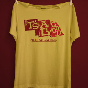The phrase 'It's All Good' inside a state shape of nebraska in the color of red with the text 'Nebraska apparel company' on a yellow maze shirt with a deep red background.