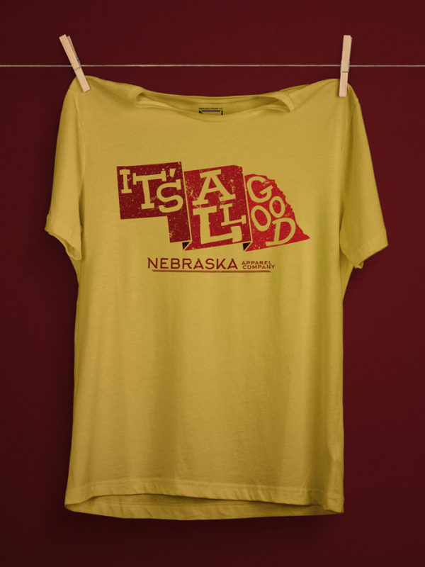The phrase 'It's All Good' inside a state shape of nebraska in the color of red with the text 'Nebraska apparel company' on a yellow maze shirt with a deep red background.
