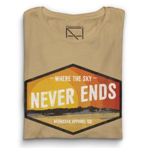 A heather Tan Shirt, folded and ready to ship. This design has a retro and grungy Nebraska landscape design that reads 'where the sky never ends' and stylized sunset. The design is contained within a hexogon black box and the shirt is on a muted red background.
