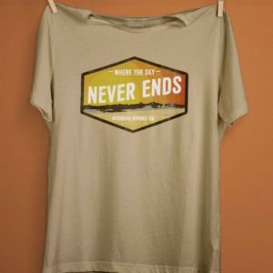 A heather Tan Shirt, hanging on a close-line to show how the shirt naturally lays. The shirt has a retro and grungy Nebraska landscape design that reads 'where the sky never ends' and stylized sunset. The design is contained within a hexogon black box and the shirt is on a muted red background.