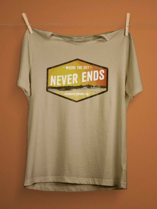 A heather Tan Shirt, hanging on a close-line to show how the shirt naturally lays. The shirt has a retro and grungy Nebraska landscape design that reads 'where the sky never ends' and stylized sunset. The design is contained within a hexogon black box and the shirt is on a muted red background.